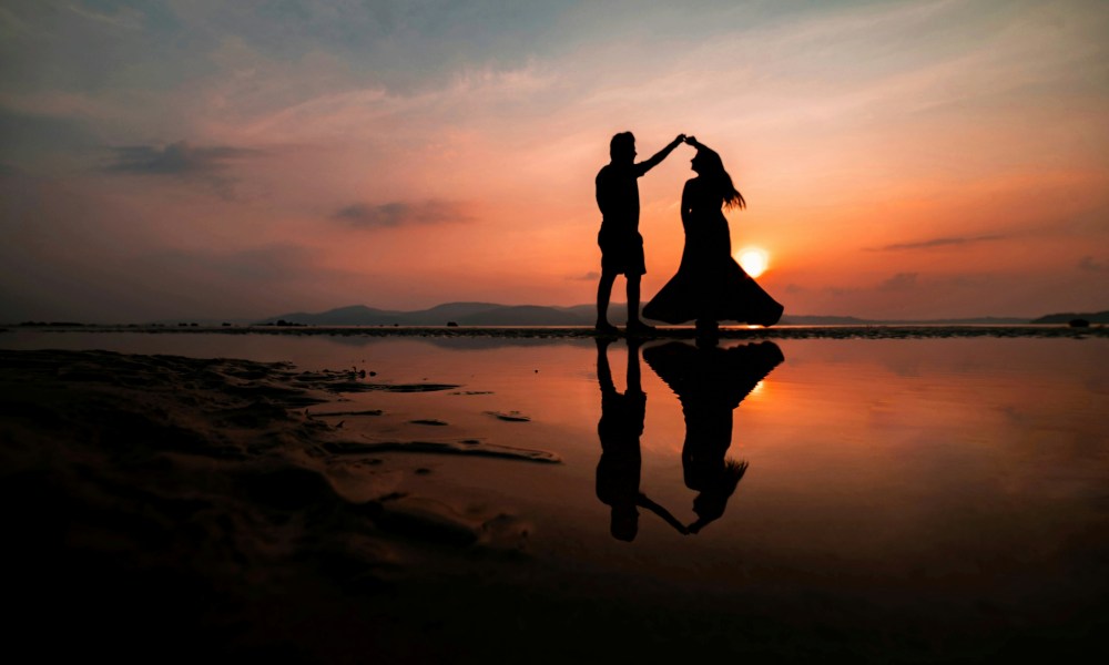 Silhouette of couple dancing by water at sunset, with reflections.